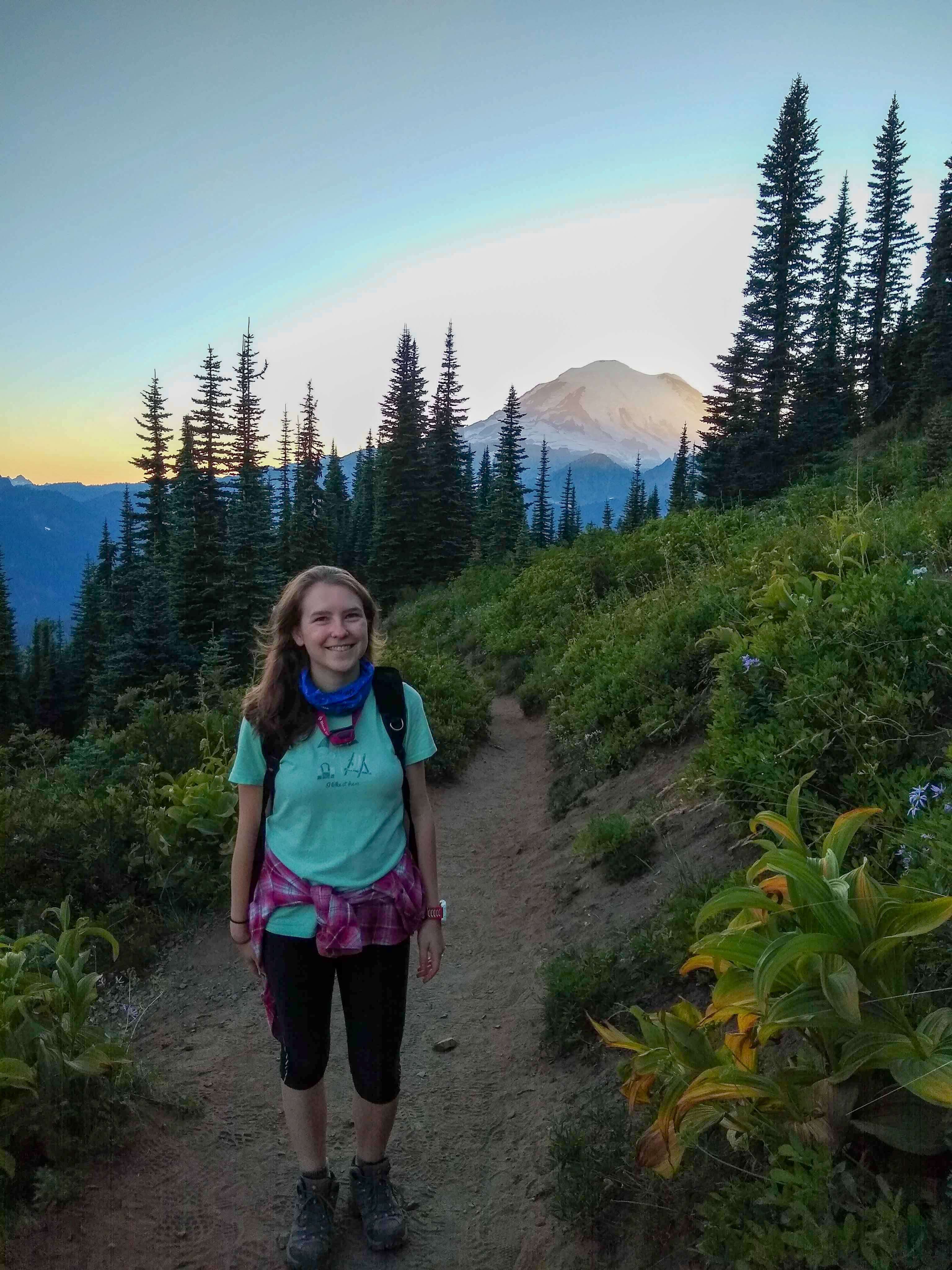 Female hiker standing on the Naches Peak Loop trail with the sun setting behind Mount Rainier in the background