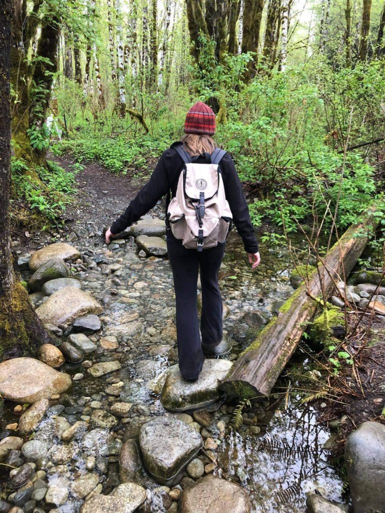 A hiker wearing a backpack walking across a stream in the woods