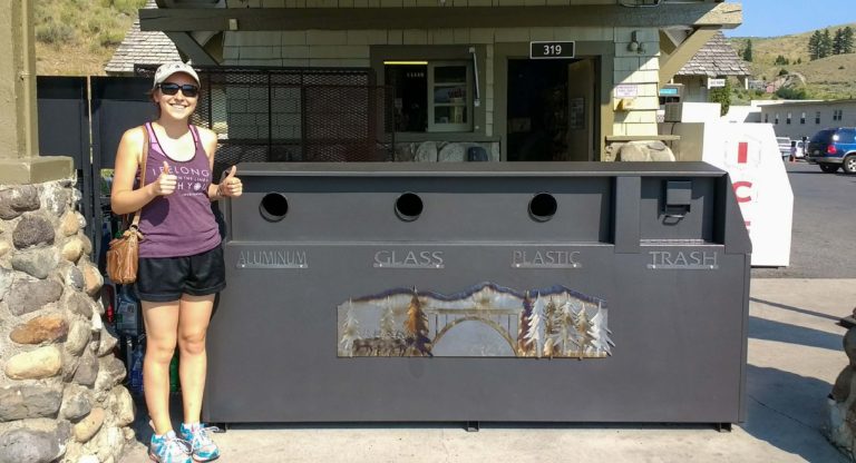 Image of a female camper giving two thumbs up next to a set of metal recycling and trash bins in a national park