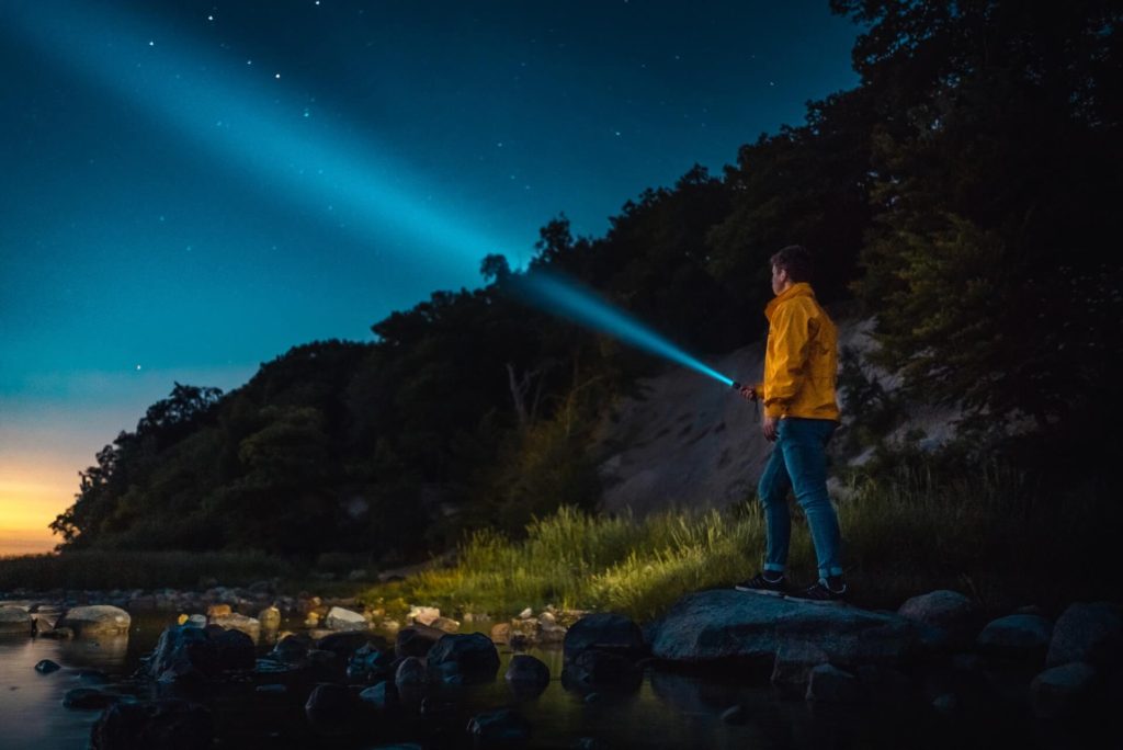 A man stands on a rock along the water while pointing a flashlight into the night sky