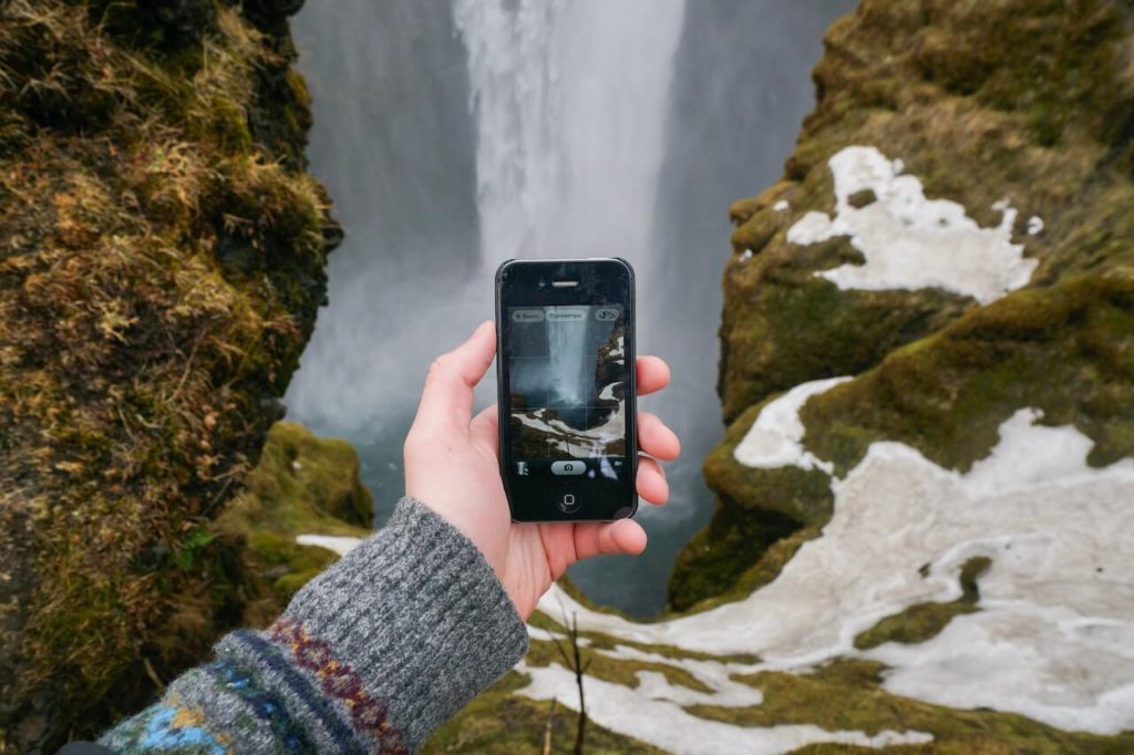 A hand holding a cell phone camera in front of a waterfall