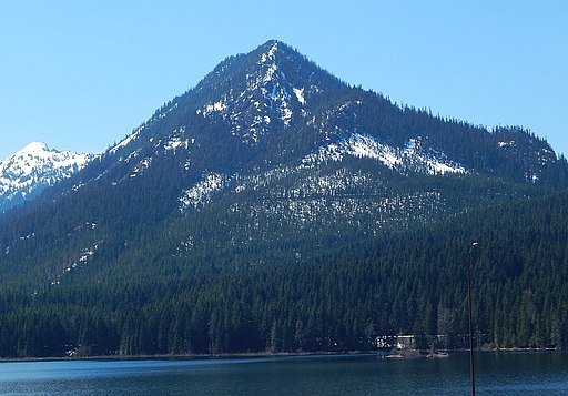 A mountain covered in green trees and spots of snow behind a lake