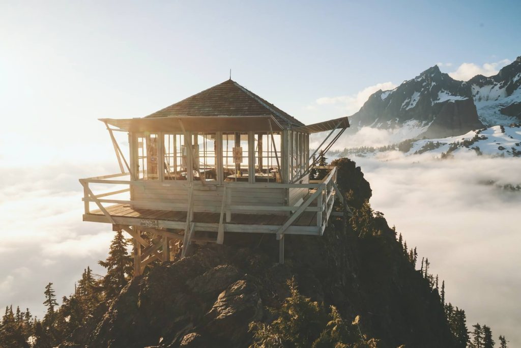 A white fire lookout stands on a rocky point above clouds.